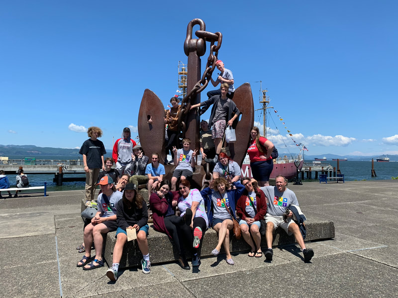 a group of youth and adults gathered in front of an anchor art installation on a pier